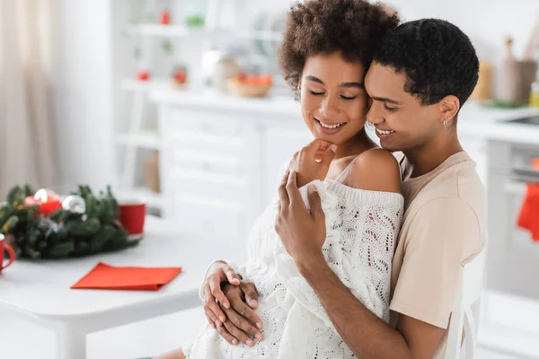 笑着的非洲裔美国男人拥抱着穿着白色针织毛衣的性感女人 接近模糊的圣诞花环 — 图库照片