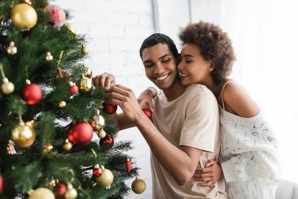 sexy african american woman embracing happy boyfriend decorating pine tree with christmas balls