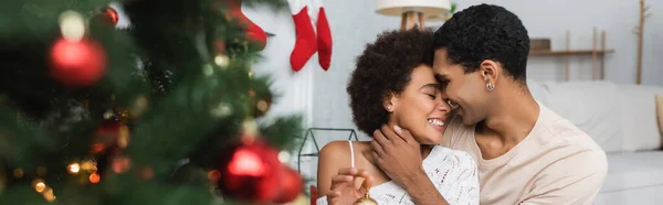 Young Happy African American Couple Embracing Blurred Christmas Tree Banner — Stock Photo, Image