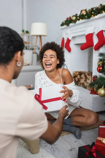 Laughing African American Woman Holding Gift Box While Sitting Floor — Stock Photo, Image