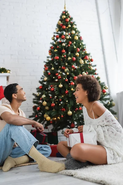 幸せなアフリカ系アメリカ人男性見ますクリスマスツリー近くセクシーガールフレンドとともにギフトボックス — ストック写真