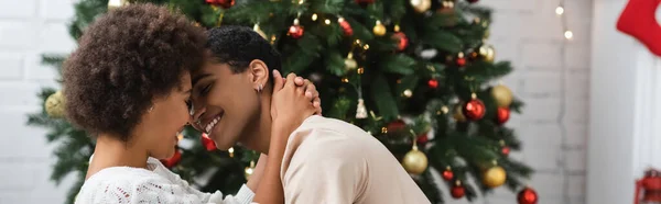 若いアフリカ系アメリカ人の女性がクリスマスツリーの近くで笑顔のボーイフレンドを抱きかかえて — ストック写真