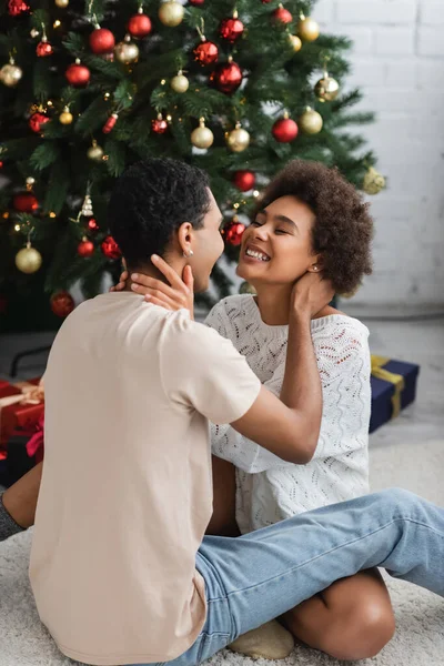 excited african american woman hugging with boyfriend while sitting on floor near christmas tree