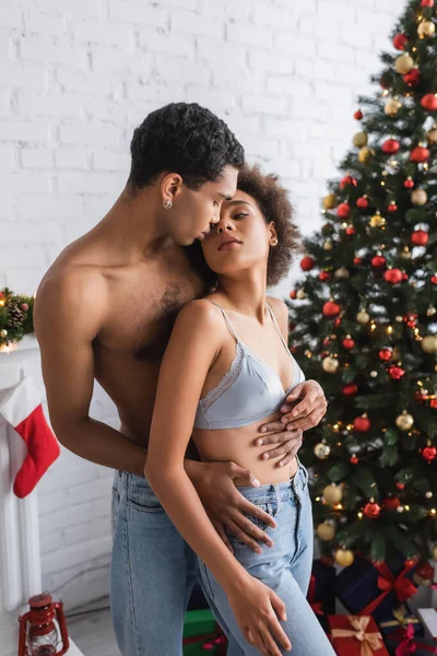 热情的非洲裔美国女人看着赤身裸体的男人拥抱着她的圣诞树 — 图库照片