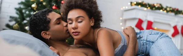 sexy african american woman in jeans and bra near young boyfriend lying on bed, banner