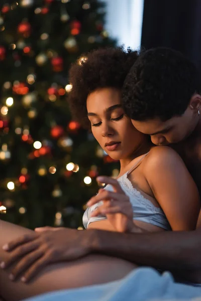 young african american man hugging and kissing sexy girlfriend in bedroom with christmas lights