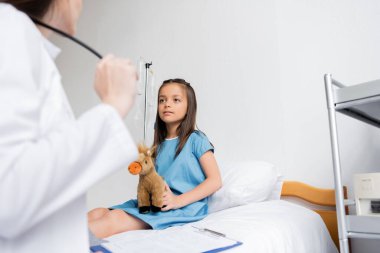 Girl holding soft toy near blurred doctor and clipboard in hospital ward  clipart