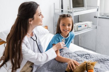Hastane yatağında, çocuğun göğsüne yakın steteskop tutan gülümseyen doktor. 