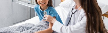 Cropped view of pediatrician holding stethoscope near chest of kid on bed in clinic, banner  clipart