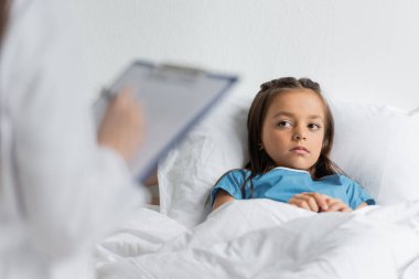 Klinikte yatan çocuk, bulanık bir doktorun yanında panoya yazı yazıyor. 