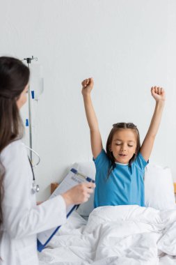 Hastane koğuşunda elinde dosyayla çocuk doktorunun yanında oturan heyecanlı çocuk. 