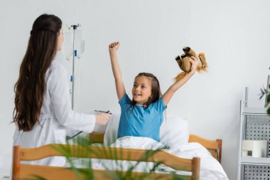 Elinde oyuncak olan çocuk hastane koğuşunda elinde dosya olan çocuk doktorunun yanında evet işareti yapıyor. 