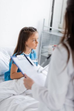 Hastane koğuşundaki bulanık çocuk doktorunun yanında yatağın üzerinde bir bardak su tutan çocuk. 