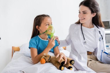 Smiling doctor with clipboard looking at happy child with oxygen mask in hospital  clipart