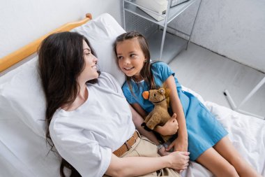 High angle view of child in patient gown holding toy and hand of mom on hospital bed  clipart