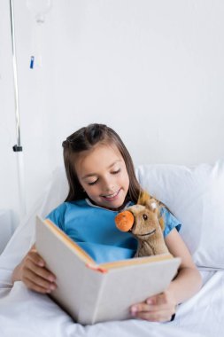 Neşeli çocuk kitap tutuyor ve klinikteki yatakta yumuşak oyuncaklara bakıyor. 