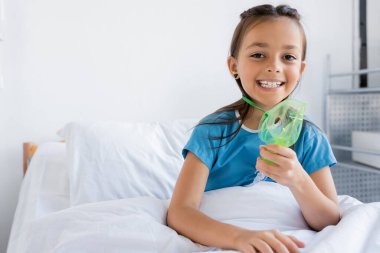 Cheerful kid holding oxygen mask and looking at camera on bed in clinic  clipart