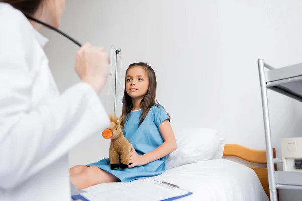 女の子保持ソフトおもちゃ近くぼやけた医師とクリップボードで病院病棟 — ストック写真