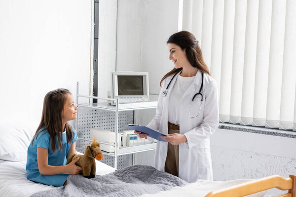 Вид сбоку улыбающегося врача с планшетом, смотрящего на пациента с мягкой игрушкой в клинике 