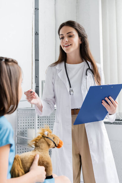 Smiling doctor holding clipboard and talking to child with toy in hospital ward 