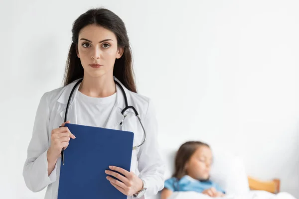 Pediatrician Holding Clipboard Looking Camera Blurred Child Clinic — Stock Photo, Image