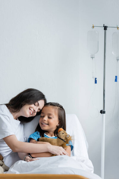 Parent touching hand of smiling child with soft toy on bed in clinic 