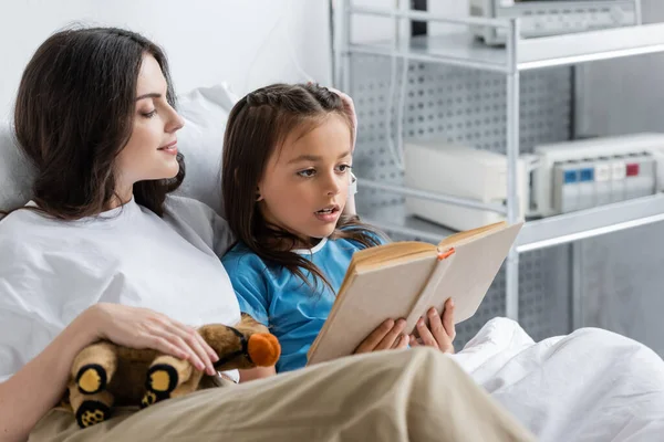 Kid reading book near mom with soft toy on hospital bed