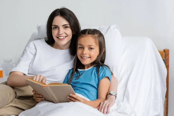 Lächelnde Frau Und Tochter Blicken Kamera Neben Buch Auf Bett — Stockfoto