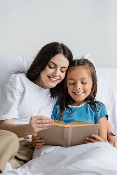 Young parent hugging daughter with book on bed in clinic