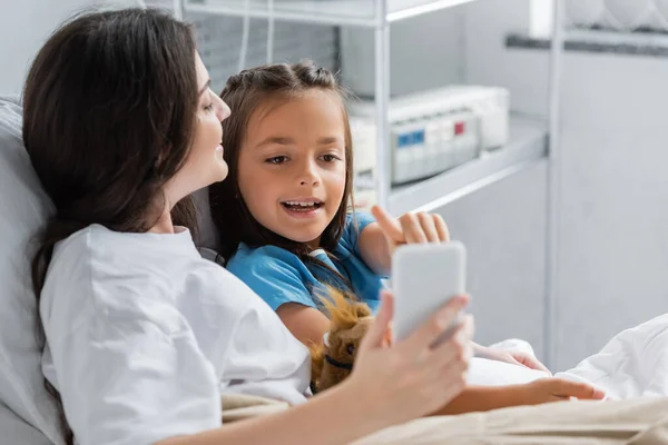 Gülümseyen Kız Klinikte Yatağında Annesinin Yanında Akıllı Telefonu Işaret Ediyor — Stok fotoğraf