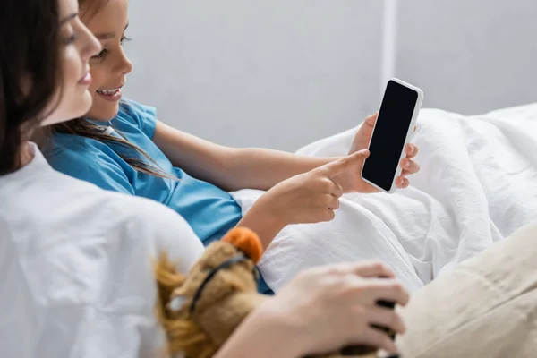 Child pointing at smartphone near blurred mother in hospital ward