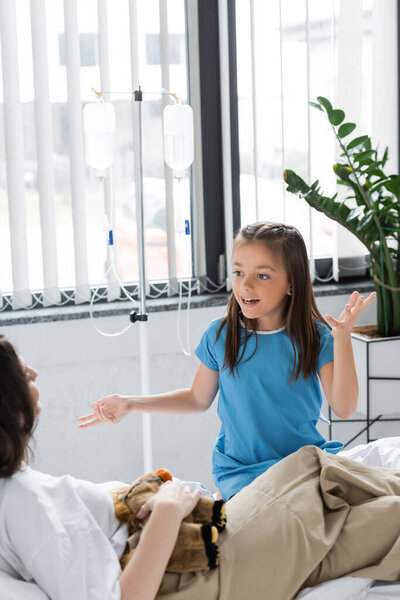 Girl talking to blurred mom near intravenous therapy in hospital ward 