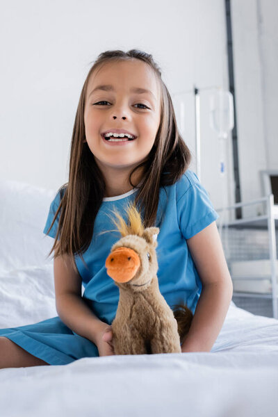 Smiling kid in patient gown looking at camera near soft toy on bed in clinic 