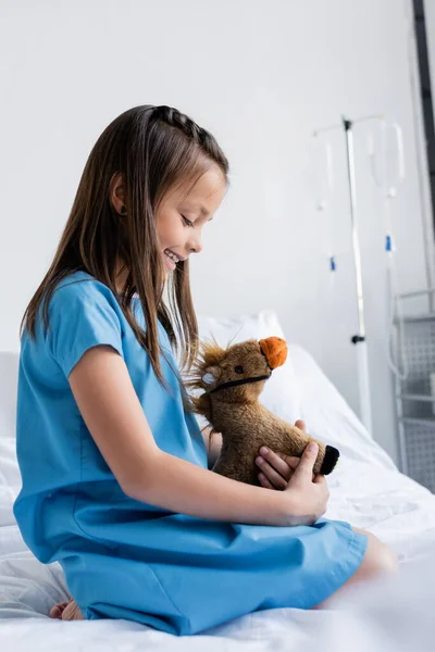 临床上穿着病人长袍抱着软玩具的儿童侧视图 — 图库照片