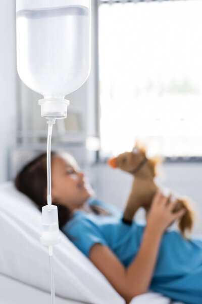 Bottle of intravenous therapy near blurred kid with toy on bed in clinic 