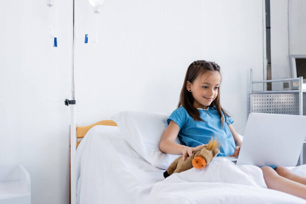 Smiling kid in patient gown holding toy and using laptop on bed in hospital 
