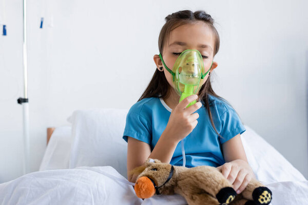 Sick child using oxygen mask near toy on bed in hospital 