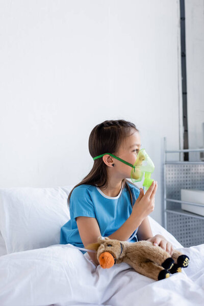 Side view of child holding oxygen mask and toy on bed in hospital ward 