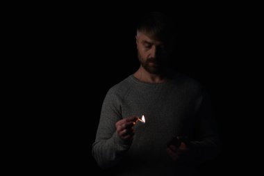 Karanlıkta elinde kibrit kutusu olan ve yanan kibrite bakan adam siyahta izole edilmiş.