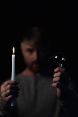 selective focus of light bulb and burning candle in hands of blurred man isolated on black clipart