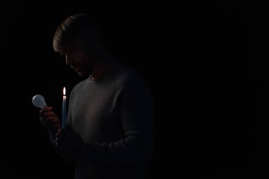 man in darkness holding lit candle and electric bulb isolated on black clipart