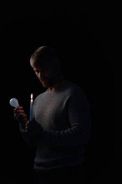 Erişkin bir adam elektrik kesintisi sırasında ampul ve mum tutuyor.