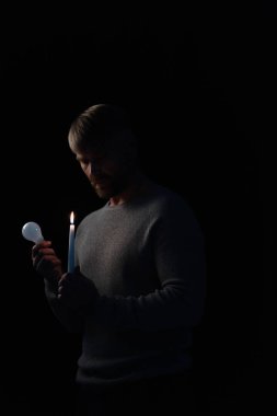 adult man holding bulb and lit candle during electricity shutdown isolated on black clipart
