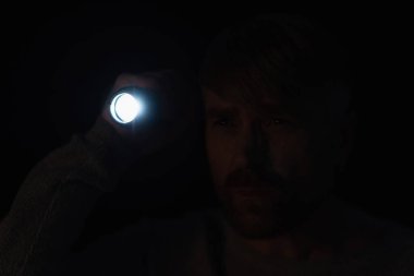 man with bright flashlight looking away in darkness isolated on black clipart