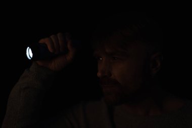 man looking away in darkness while holding bright flashlight isolated on black clipart