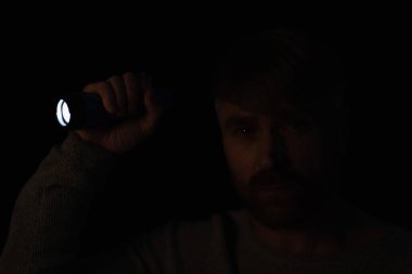 man in darkness looking at camera while holding bright flashlight isolated on black clipart