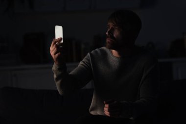 Karanlık mutfakta cep telefonuyla oturan gergin adam güç kesintisi sırasında bağlantı arıyor.