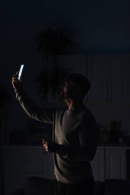Adam karanlık mutfakta elinde akıllı telefonuyla dikiliyor ve elektrik kesintisi sırasında bağlantı arıyor.