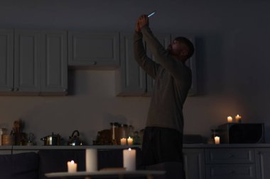 man holding cellphone in raised hands while catching mobile signal in dark kitchen near burning candles clipart