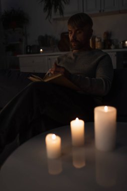 man reading book during electricity shutdown near lit candles in dark kitchen clipart